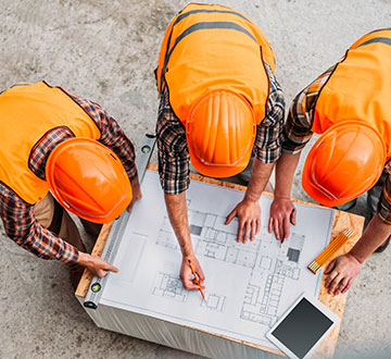 Três construtores de uniformes e capacetes laranja analisando a planta da obra.