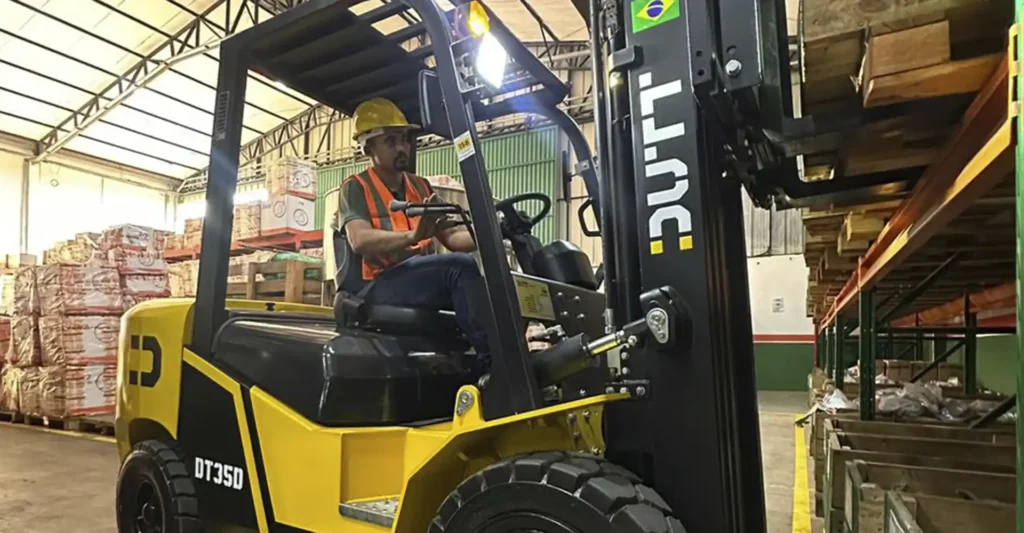 Empilhadeira a combustão Dutt DT35D amarela em operação dentro de armazém, transportando pallet de madeira em estantes metálicas de alta capacidade.